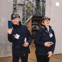 Zwei Menschen mit Schutzhelmen stehen vor der DAM Baustelle und lachen